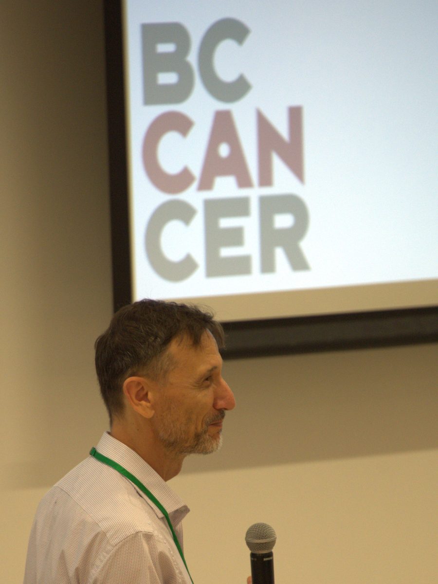Dr. Inan&ccedil; Birol speaking with a microphone, a BC Cancer slide on the screen behind him